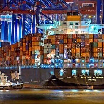 A brightly lit cargo ship at Hamburg harbor with stacked containers and a tugboat.